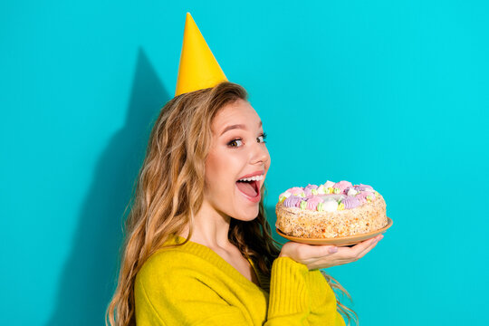 Young woman with blonde hair wearing a yellow party hat holds a colorful cake against a bright blue background for a festive birthday promotion and celebration