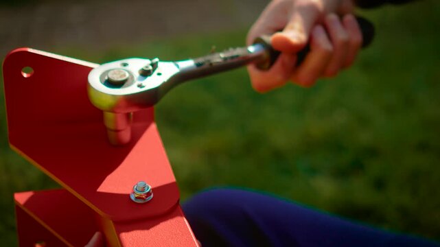 Macro view wrench gripping bolt head on red metal structure during installation. Craft repair activity with hardware and tool kit on grass background. Maintenance concept highlighting close detail of