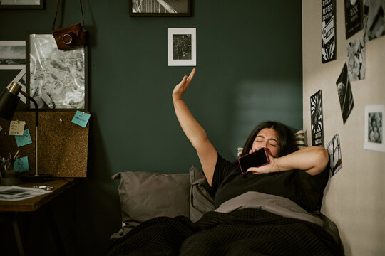 Young biracial woman waking up, stretching in bed and holding smartphone while yawning. Ideal visual asset for sleep tracking app advertising, digital wellbeing content