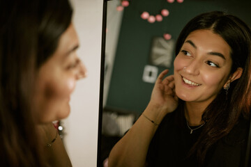 Young biracial woman smiling while looking at mirror reflection, checking skin and hair. Visual for...