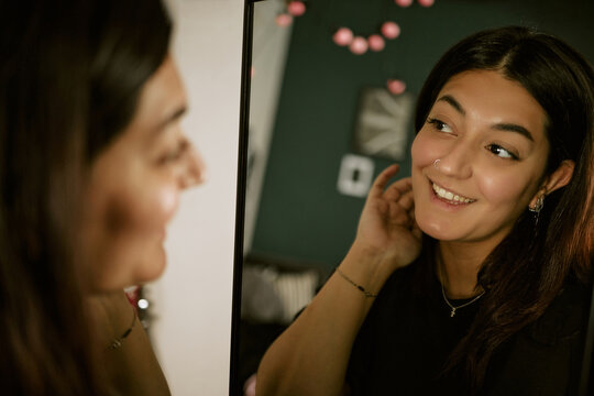 Young biracial woman smiling while looking at mirror reflection, checking skin and hair. Visual for skincare routines, beauty marketing, personal care branding, closeup shot