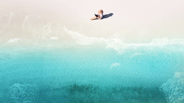 Aerial view of woman lying with laptop on pink sand beach. Seamless looping drone shot of remote work and digital nomad lifestyle by turquoise ocean waves