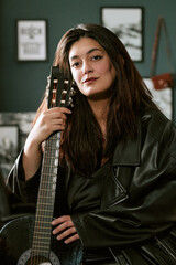 Portrait of young biracial woman holding acoustic guitar indoors, looking at camera. Suitable for...