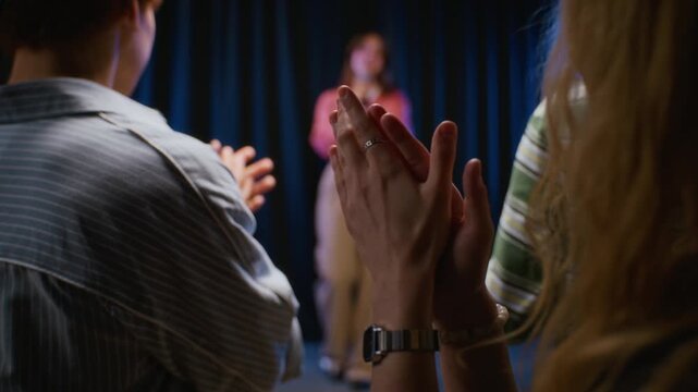 Caucasian young woman speaking into microphone on stage as classmates applaud from over shoulder view. Ideal for promoting youth theater workshops, acting classes and public speaking courses