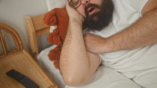Man with beard and glasses sleeping with head on hand on bed in building, orange tassel pillow visible; quiet calm.