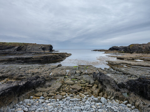 Grey skies loom over rocky shoreline. Weathered stones form natural pathways through tide pools. Ripples in water suggest gentle sea movement. Foggy air adds serene, isolated atmosphere