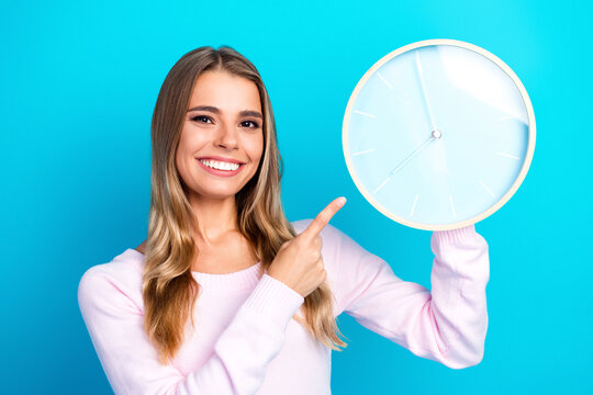 Smiling young woman pointing at wall clock on bright blue background to show time, punctuality and reminder for schedule or deadline