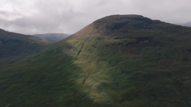 Aerial view of a rounded grassy mountain slope with patches of light and shadow beneath a moody sky.
