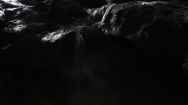 Dark, moody close-up of water cascading over wet, textured rocks under dramatic lighting