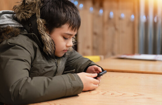 Child using smartphone at wooden table in winter clothing, focused on mobile screen indoors, digital technology lifestyle for autumn, winter, back to school, education and communication