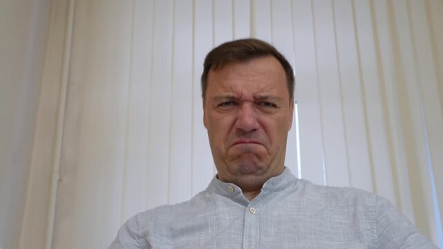 Distressed man crying and expressing sadness in front of window blinds.