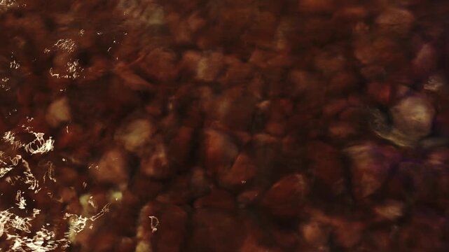 A dark, abstract, high-angle shot capturing brown riverbed stones under shallow, flowing water with sparkling surface reflections and natural light