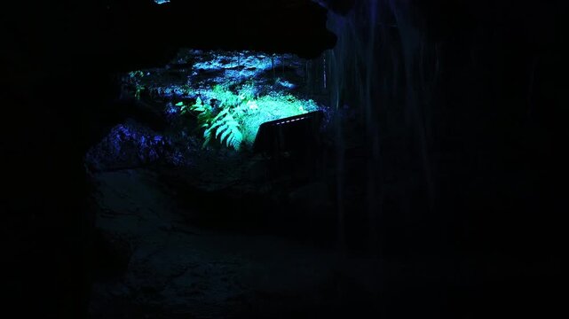 Green illuminated cave interior with ferns and a small waterfall, dark and moody