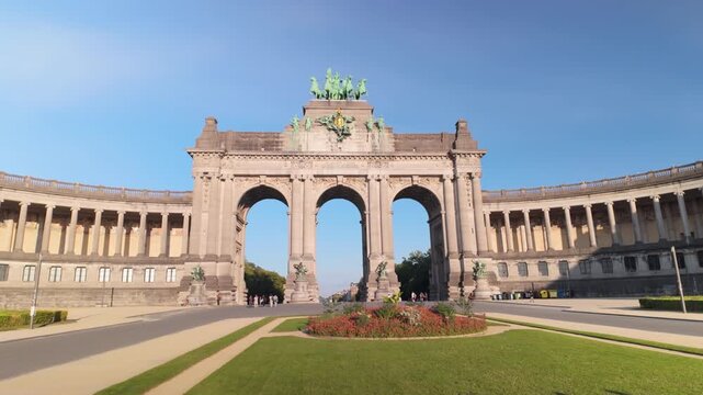 Historic triumphal arch and colonnade at Cinquantenaire park in Brussels, Belgium, framed by lawns, flower beds, and a clear blue sky.