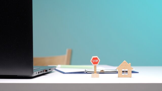 Miniature house and stop sign resting on a desk with a laptop and documents, symbolizing a halt or caution in real estate transactions, property investment, or mortgage processes
