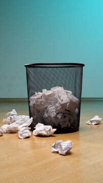 Crumpled paper filling a mesh waste bin and scattered on the wooden floor, representing discarded ideas, frustration, failure, and the creative process of trial and error in an office setting
