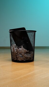 Black metal mesh wastebasket on a wooden floor filled with crumpled paper and a discarded electronic tablet, symbolizing obsolescence and the need for e waste recycling