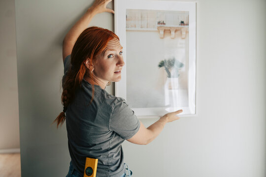 Redhead woman looking over shoulder while hanging framed picture on wall in new apartment