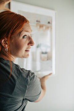 Close-up of redhead woman hanging painting on wall in her new home