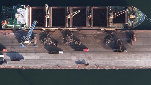 Bulk cargo ship unloading dark material beside quay at Port of Veracruz, aerial, excavator digger transports from pile to trucks
