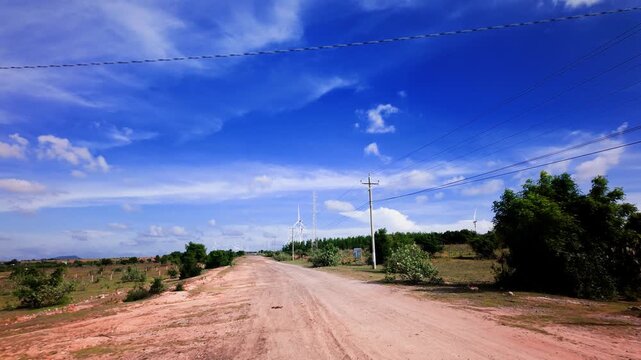 The view shows a dirt road in Hoa Thang, Lam Dong. Trees line the sides while the sky is clear. This area features open fields and simple country life during daylight hours.