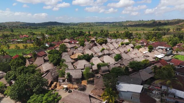 Aerial view of Sade Traditional Village in Lombok, Indonesia, showing dense Sasak thatched-roof houses surrounded by tropical farmland and rolling green hills.