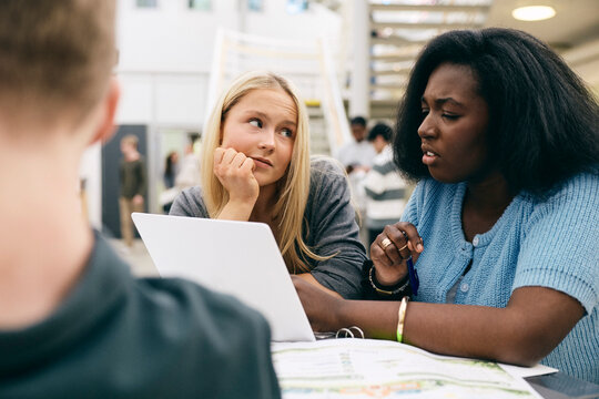 Blond girl listening to female friend sharing ideas while using laptop in high school