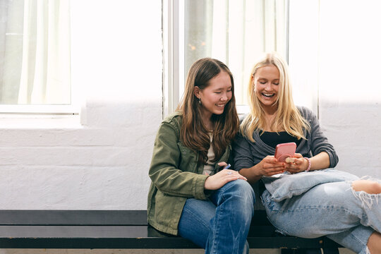 Happy multiracial teenage girls spending their leisure time using smart phone in college lobby