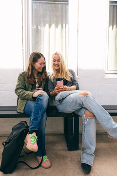 Full length of happy multiracial teenage girls spending their leisure time using smart phone while sitting on bench in c