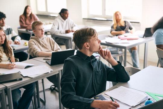 Focused teenage boy sitting with books at desk while leaning on elbow in classroom at college