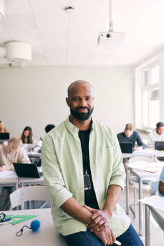 Portrait of smiling mature male high school teacher with shaved head sitting at desk in classroom