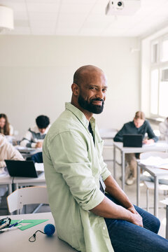 Portrait of smiling male high school professor with shaved head sitting at desk in classroom