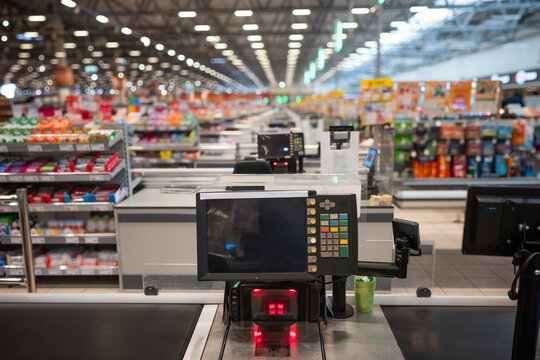 Modern supermarket checkout terminal with screen, barcode scanner, red light, and conveyor belt in busy store aisle with shelves and products, retail shopping concept