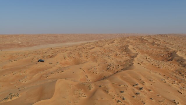 Aerial view of the vast orange sand dunes of Wahiba Sands with a 4x4 vehicle parked on the undulating terrain under a clear blue sky in Muscat, Muscat Governorate, Oman.