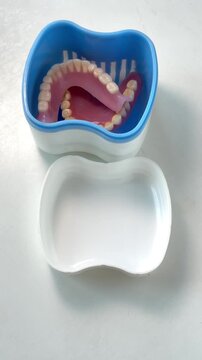 An elderly person uses a blue toothbrush to clean a removable denture inside a plastic container on a windowsill.