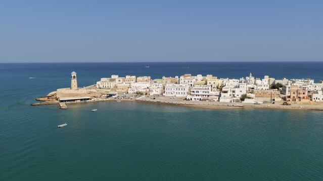 Aerial view of Al Ayjah Lighthouse and the white buildings of the historic coastal town of Sur, situated along the turquoise waters of the Gulf of Oman. Sur, Ash Sharqiyah South G, Oman.