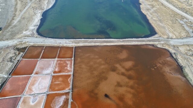 Aerial view of colorful salt evaporation ponds with varying shades of brown and red, separated by earthen dikes, adjacent to a turquoise body of water in Qurayyat, Muscat Governorate, Oman.