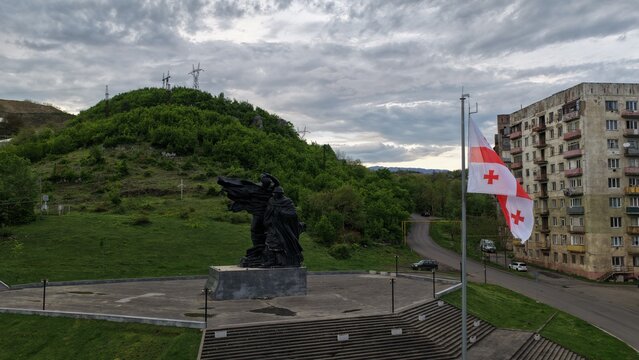 Aerial view of the monument with the Georgian flag flying nearby, set against a backdrop of green hills and an apartment building in Chiatura, Imereti, Georgia.