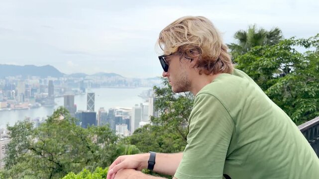 Young Blond Man Using Smartphone and Photographing with View of Hong Kong Skyscrapers from Above &ndash; Urban Panorama with City Skyline