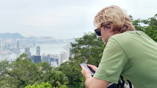 Young Blond Man Using Smartphone and Photographing with View of Hong Kong Skyscrapers from Above &ndash; Urban Panorama with City Skyline
