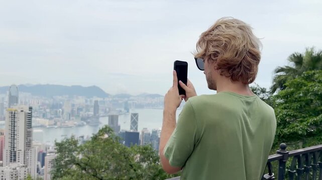 Young Blond Man Using Smartphone and Photographing with View of Hong Kong Skyscrapers from Above &ndash; Urban Panorama with City Skyline