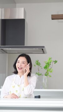 A woman in her 50s&mdash;perhaps a homemaker&mdash;smiling as she makes a call on her smartphone at home; a lifestyle where she chats with family and friends or asks questions and makes inquiries in between house