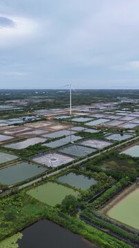 An aerial view of wind turbines for electricity generation near the sea with shrimp ponds and farmland below captured by a drone camera.