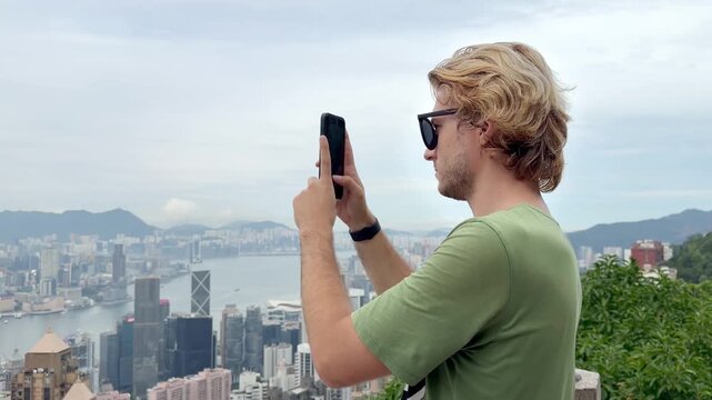 Young Blond Man Using Smartphone and Photographing with View of Hong Kong Skyscrapers from Above &ndash; Urban Panorama with City Skyline