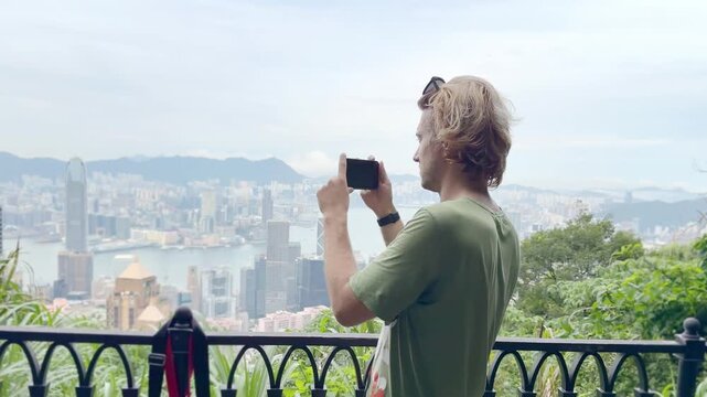 Young Blond Man Using Smartphone and Photographing with View of Hong Kong Skyscrapers from Above &ndash; Urban Panorama with City Skyline