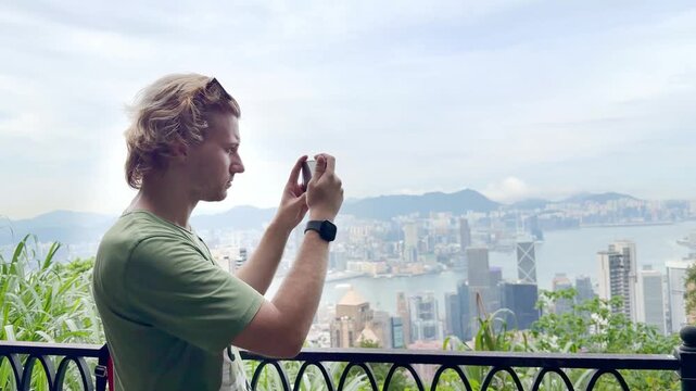 Young Blond Man Using Smartphone and Photographing with View of Hong Kong Skyscrapers from Above &ndash; Urban Panorama with City Skyline