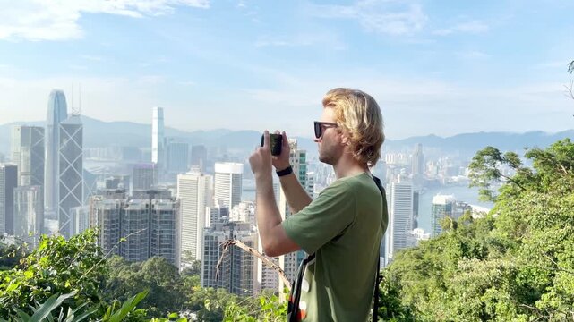 Young Blond Man Using Smartphone and Photographing with View of Hong Kong Skyscrapers from Above &ndash; Urban Panorama with City Skyline
