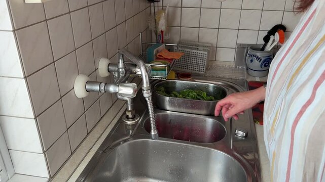 Close-up of an unrecognizable woman trimming the thin roots from freshly washed red radishes at a kitchen sink, with clean radish leaves lying nearby during food preparation, static shot