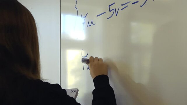 Female student writing a system of two linear equations with two variables on a white dry erase board with a blue marker, demonstrating her mathematical problem solving skills in a classroom