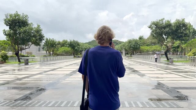 Young Blond Man Walking on Wide Park Path in Shenzhen and Talking on Smartphone &ndash; Back View with Tropical Greenery and Blue Sky